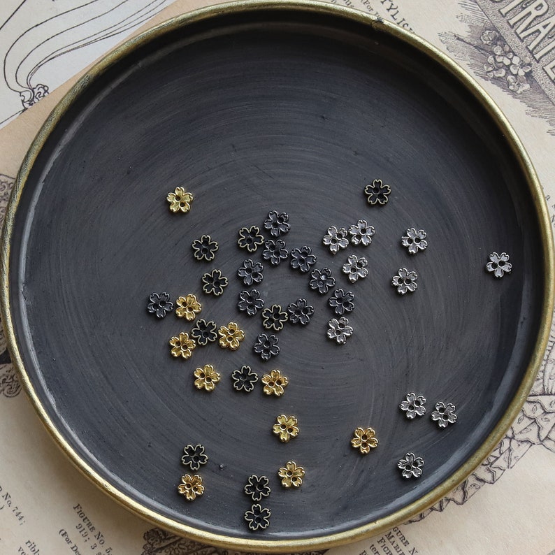 May include: A collection of small metal flower charms in gold, silver, and black colors. The charms are scattered on a dark gray surface.