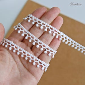 Pode incluir: Fita de renda branca com borda ondulada e pequenas bolinhas redondas. A fita é disposta em um padrão em camadas, com o texto "Charlene" no canto superior direito. Adequada para vários projetos de artesanato.