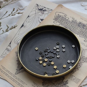 May include: A black dish with a gold rim filled with small silver and gold flower-shaped beads.