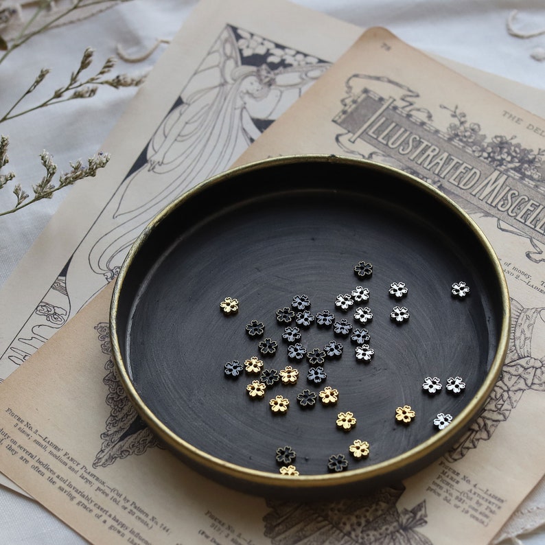 May include: A black dish with a gold rim filled with small silver and gold flower-shaped beads.