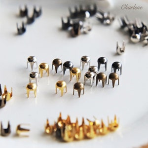 Puede incluir: Surtido de tachuelas metálicas en dorado, plateado y negro. Las tachuelas tienen una parte superior redonda y tres puntas. Las tachuelas están dispuestas sobre una superficie blanca.