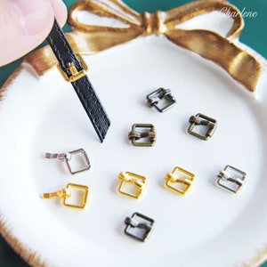 May include: Assortment of metal buckles in silver, gold, and bronze finishes. The buckles are square shaped with a small loop on one side. A black leather strap with a gold buckle is also included.
