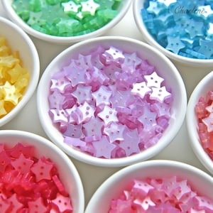 May include: A collection of small, star-shaped buttons with two holes in the centre, organised by colour in white bowls. The buttons feature a pearlescent finish and are available in shades of yellow, green, blue, purple, pink, and red.