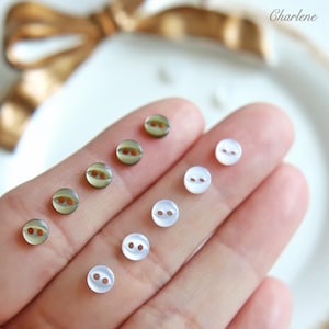 May include: A collection of small, round buttons in two distinct styles. Some are a translucent, pale green with a reddish-brown center, while others are white and pearlescent. The buttons have two holes for sewing.