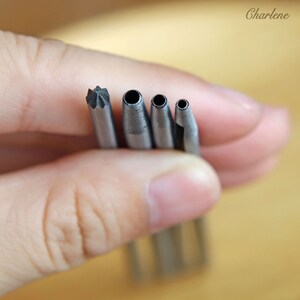 以下が含まれることがあります： 手で持たれた4つの金属製穴あけ工具のセット。工具は、星型のパンチと、サイズが小さくなる3つの円形パンチを含む、さまざまな先端デザインを持っています。工具は銀色です。