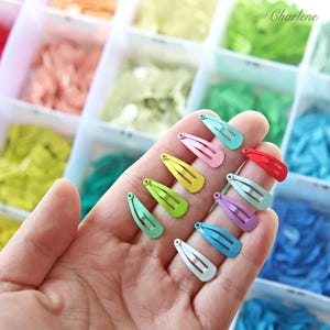 Könnte beinhalten: Eine Hand hält eine Sammlung kleiner, farbenfroher Haarspangen aus Metall. Die Spangen sind in verschiedenen Farben erhältlich, darunter Rot, Rosa, Grün, Gelb, Blau und Lila. Der Hintergrund zeigt einen Aufbewahrungsbehälter mit Fächern, die mit farbenfrohen Materialien gefüllt sind.