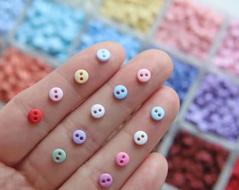 Op de afbeelding: Een verzameling kleine, kleurrijke knopen in verschillende pasteltinten, waaronder rood, roze, geel, groen, blauw en paars. De knopen zijn rond met twee gaatjes en worden op een hand getoond. Het woord "Charlene" is zichtbaar.