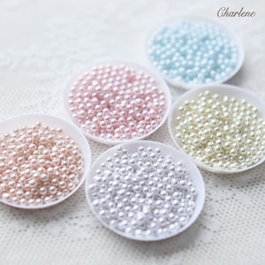 Puede incluir: Cinco pequeños cuencos blancos llenos de cuentas de perlas en varios colores: blanco, rosa claro, crema y azul claro.