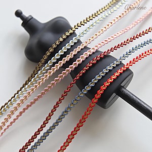 May include: A collection of six decorative braided trims featuring a metallic gold center cord flanked by colored edges in brown, light blue, pink, dark red, blue, and red. The trim is displayed over a black mannequin torso with the text Charlene.