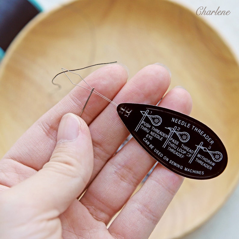May include: A black plastic needle threader with instructions on how to use it. The threader is being held in a hand, and a needle and thread are being threaded through it.