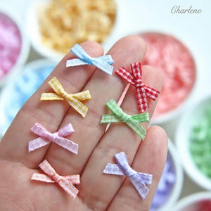 May include: A collection of seven small fabric bows in various pastel and bright colors, each featuring a delicate gingham check pattern. The bows are arranged in a cluster, showcasing light blue, yellow, pink, orange, red, green, and purple.