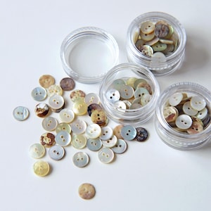 May include: A collection of small, round, two-hole buttons made of iridescent mother-of-pearl. The buttons feature varied natural tones of cream, beige, and brown. Some buttons are loose, while others are stored in three clear plastic jars.