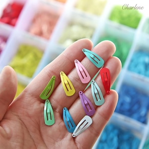 Op de afbeelding: Een verzameling kleurrijke metalen haarclips in verschillende tinten, waaronder groen, geel, roze, blauw en rood. De clips worden in een hand gehouden, met een vage achtergrond van andere kleurrijke items. Het woord "Charlene" is zichtbaar.