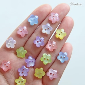 Könnte beinhalten: Eine Sammlung von blütenförmigen Knöpfen in verschiedenen Pastellfarben, darunter Rosa, Blau, Gelb, Grün und Lila. Jeder Knopf hat zwei Löcher zum Annähen. Der Name "Charlene" ist oben rechts zu sehen.