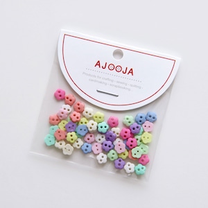 May include: A clear plastic bag filled with small, colorful flower-shaped buttons. The buttons are in shades of pink, purple, blue, green, and yellow. The bag has a white label with the text "AJOOJJA Products for crafting, sewing, quilting, cardmaking, scrapbooking..."