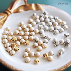 May include: A white dish containing three groups of pearl-like buttons. The buttons are arranged in a circular pattern. The buttons are gold, silver, and white.