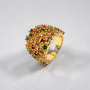 Könnte beinhalten: Ein breiter goldfarbener Ring mit einem durchbrochenen, texturierten Metalldesign, besetzt mit zahlreichen kleinen, mehrfarbigen Edelsteinen, darunter rote, grüne, violette und klare Steine, die über die Oberfläche des Rings verteilt sind.