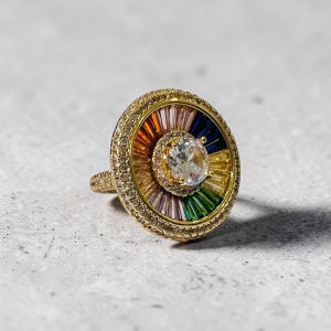 Könnte beinhalten: Ein goldener Ring mit einer großen, runden Front, die einen zentralen, klaren Stein enthält, umgeben von einem strahlenförmigen Muster aus baguettegeschliffenen, bunten Steinen in Orange, Blau, Gelb, Grün und Violett, eingefasst von kleinen Steinen.