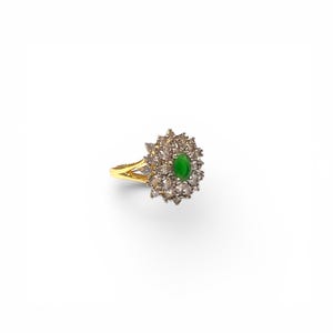 Op de afbeelding: Een gouden ring met een centrale ovaalvormige groene steen, omringd door een cluster van kleine, heldere, marquise-geslepen stenen in een bloemmotief. De ringband is verdeeld in twee dunne, gepolijste gouden segmenten die aan de basis samenkomen.