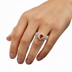 May include: A ring featuring a central oval-cut red gemstone set in a gold-toned bezel, surrounded by a halo of small clear stones. The band is crafted from a silver-toned metal and is embellished with additional clear stones along the sides.