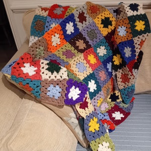May include: A colourful crocheted blanket with a patchwork design featuring squares in various colours, including red, blue, green, yellow, and white. The blanket is draped over a beige sofa.