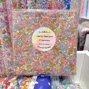 May include: A stack of Liberty Tana Lawn fabric squares in a 5-inch charm pack. The top square features a dense floral pattern in pink, yellow, and blue. A circular label reads: Liberty Tana Lawn, 5” Charm Pack, 25 pcs No Repeats, Polka Dot Secrets.