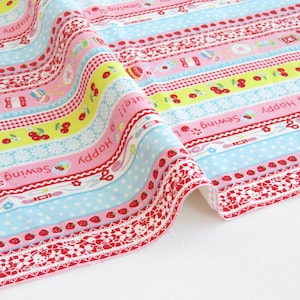 以下が含まれることがあります： ピンク、青、黄色の横縞模様のカラフルな生地。縞模様には、さくらんぼ、イチゴ、裁縫関連のアイコンのパターンがあります。生地には白いレースのトリムがあり、「Happy」、「Cute!」、「Sewing Love」の文字が見えます。