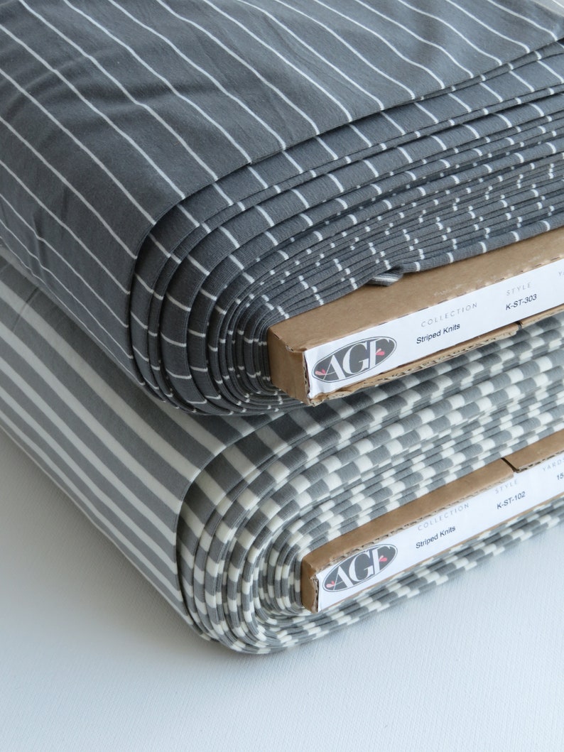 May include: Two rolls of gray and white striped knit fabric. The fabric is folded and stacked on top of each other. The fabric is labeled "Striped Knits" and "AGF" on the packaging.