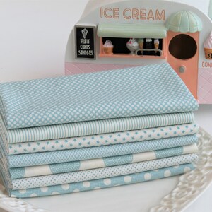 May include: A stack of eight folded fabric squares in various patterns, including polka dots, stripes, and stars, in shades of light blue and white. The fabrics are arranged on a white, decorative plate. An ice cream cart model is in the background.