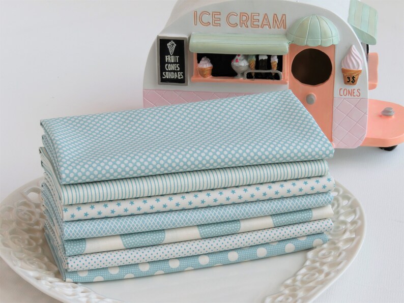 May include: A stack of eight folded fabric squares in various patterns, including polka dots, stripes, and stars, in shades of light blue and white. The fabrics are arranged on a white, decorative plate. An ice cream cart model is in the background.