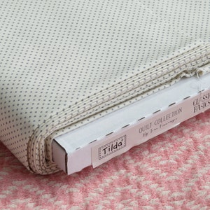 May include: A roll of off-white fabric with a repeating pattern of small, gray dots. The fabric is part of the "Classic Basics" quilt collection by Tilda. The fabric is in a cardboard box, and the background is a pink and white patterned fabric.