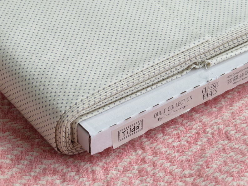 May include: A roll of off-white fabric with a repeating pattern of small, gray dots. The fabric is part of the "Classic Basics" quilt collection by Tilda. The fabric is in a cardboard box, and the background is a pink and white patterned fabric.