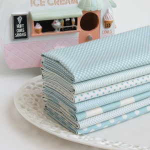May include: A stack of light blue fabric squares with various white patterns, including dots, stars, and stripes, rests on a white decorative plate. An ice cream shop figurine is in the background.