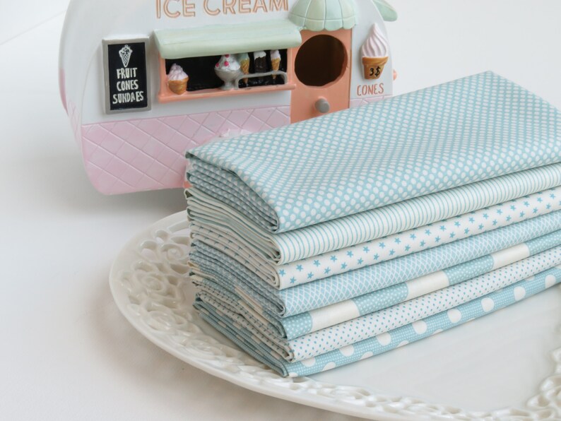 May include: A stack of light blue fabric squares with various white patterns, including dots, stars, and stripes, rests on a white decorative plate. An ice cream shop figurine is in the background.