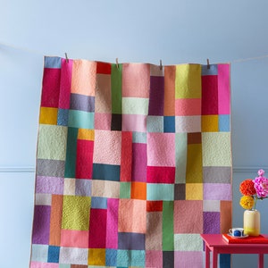 Op de afbeelding: Een kleurrijk patchwork-deken met vierkanten in verschillende kleuren, waaronder roze, rood, groen, blauw, geel en oranje. Het deken hangt aan een waslijn tegen een lichtblauwe muur.