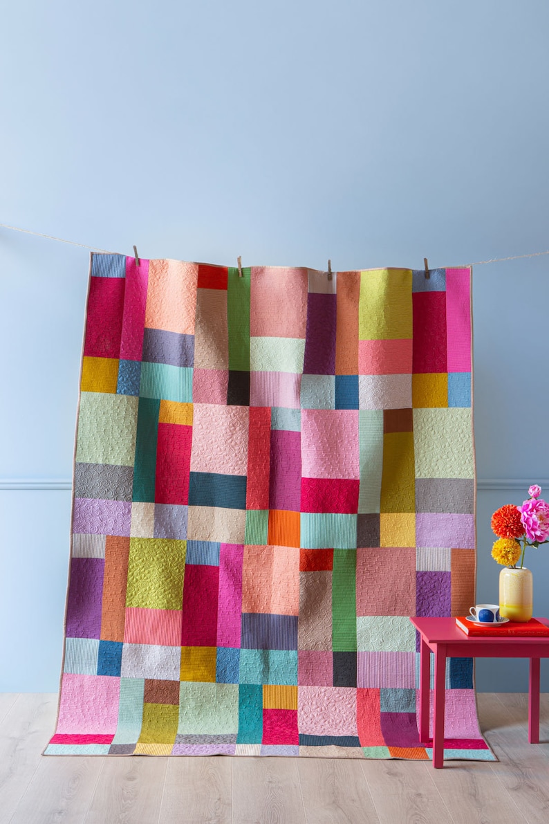 Op de afbeelding: Een kleurrijk patchwork-deken met vierkanten in verschillende kleuren, waaronder roze, rood, groen, blauw, geel en oranje. Het deken hangt aan een waslijn tegen een lichtblauwe muur.