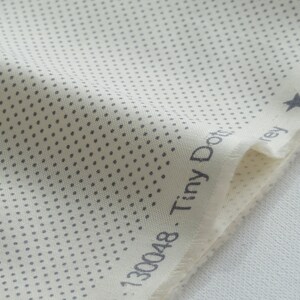 May include: Close-up of a cream-colored fabric with a pattern of tiny gray dots. The fabric has a raw edge and the words "Tiny Dots Grey" are printed on the edge. A small gray star is also visible.