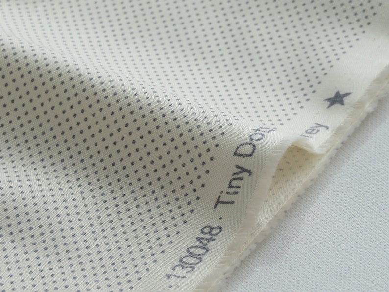 May include: Close-up of a cream-colored fabric with a pattern of tiny gray dots. The fabric has a raw edge and the words "Tiny Dots Grey" are printed on the edge. A small gray star is also visible.