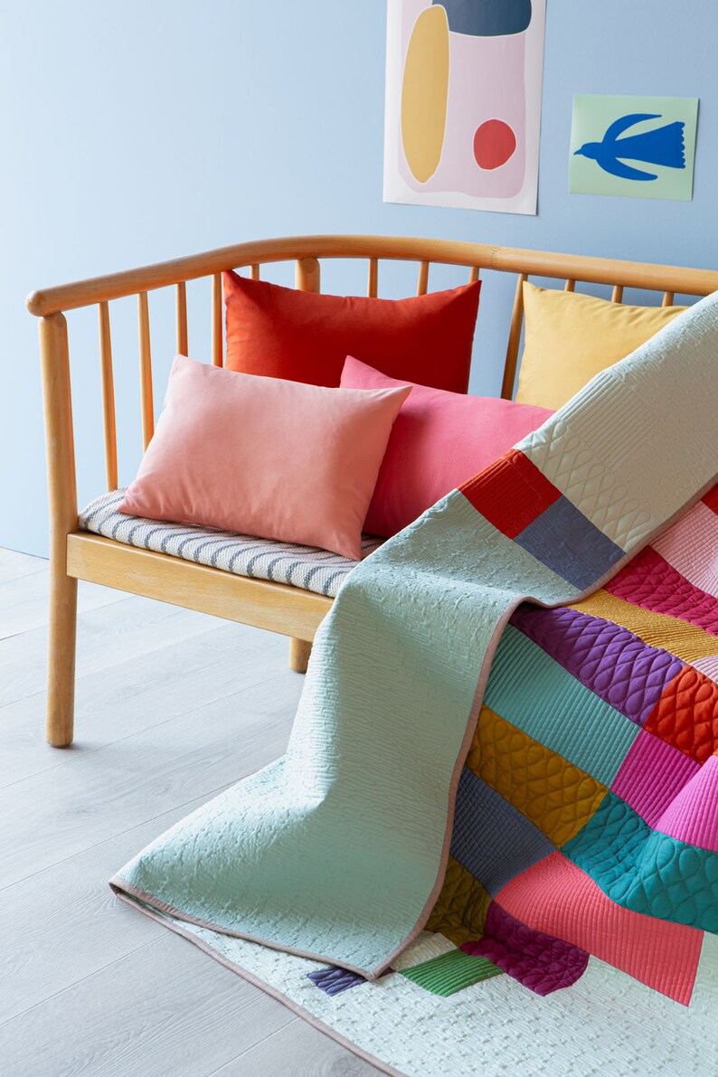 Op de afbeelding: Een lichtblauwe en witte quilt met kleurrijke patchwork-vierkanten in tinten roze, rood, paars, turquoise en geel. De quilt is gedrapeerd over een houten dagbed met gestreepte kussens en kissen in tinten roze, rood en geel.