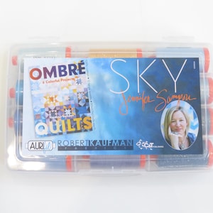 Pode incluir: Uma caixa de plástico transparente com bobinas de linha. A caixa apresenta um rótulo com o texto "Ombre Quilts" e "Sky Jennifer Sampou". As bobinas têm detalhes vermelhos e azuis.