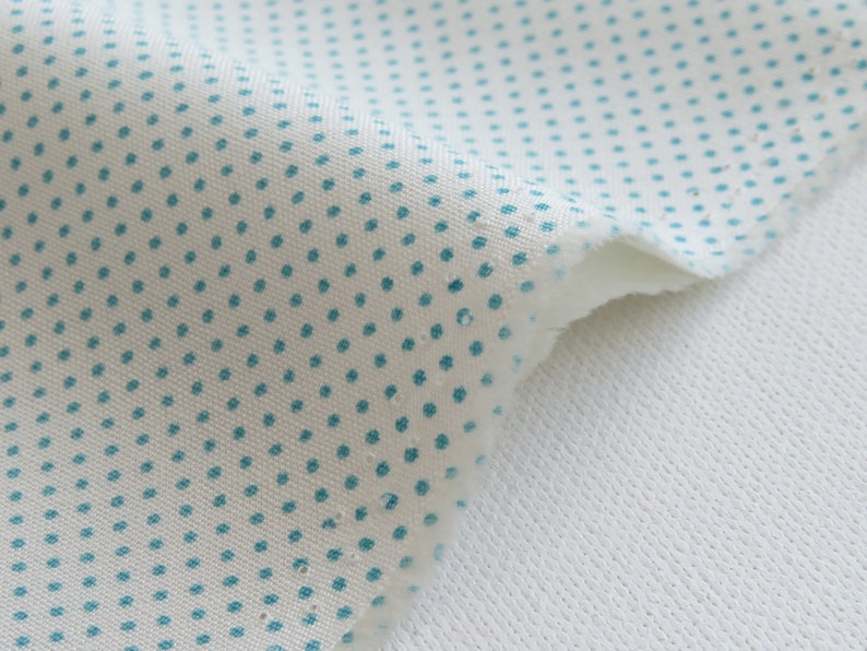 May include: Close-up of a piece of white fabric with a pattern of small, evenly spaced teal dots. The fabric is layered over a white, textured material, creating a contrast in textures. The edges of the fabric are frayed.