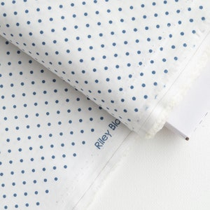 May include: White fabric with a pattern of blue polka dots. The fabric is folded over a white surface. The text "Riley Blake" is printed on the fabric.