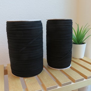 Puede incluir: Dos carretes de cord&oacute;n el&aacute;stico negro, enrollados apretados y apilados sobre una superficie de madera.
