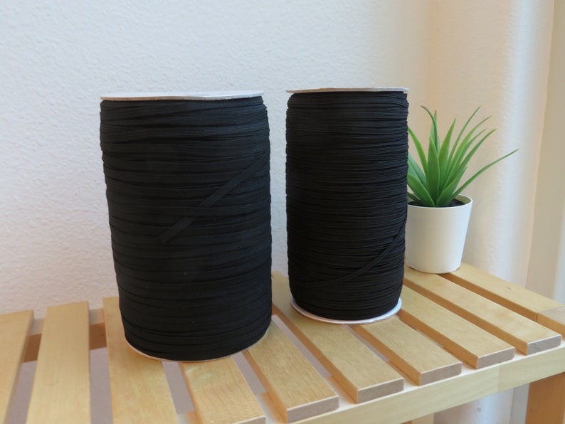 Puede incluir: Dos carretes de cord&oacute;n el&aacute;stico negro, enrollados apretados y apilados sobre una superficie de madera.