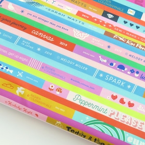 以下が含まれることがあります： ピンク、青、緑、黄色など、さまざまな色の斜めのストライプが入ったカラフルな生地。ストライプには「canvas」、「spark」、「Peppermint PLEASE」などの文字が含まれています。生地は陽気で芸術的なデザインです。