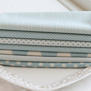 May include: A stack of folded fabric squares in various patterns, including stripes, polka dots, stars, and geometric designs. The fabrics are primarily light blue and white, arranged on a white, decorative plate. Ideal for quilting or crafting projects.