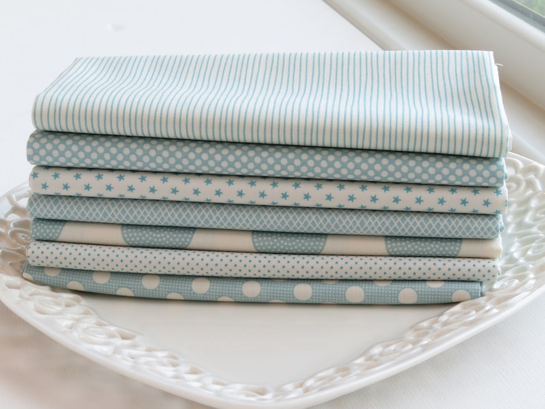 May include: A stack of folded fabric squares in various patterns, including stripes, polka dots, stars, and geometric designs. The fabrics are primarily light blue and white, arranged on a white, decorative plate. Ideal for quilting or crafting projects.