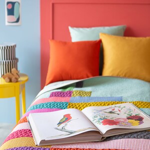 Op de afbeelding: Een kleurrijk patchwork-deken met een open boek erop. Het deken is gemaakt van verschillende gekleurde vierkanten met verschillende texturen. Het boek heeft een afbeelding van een vogel op de omslag.