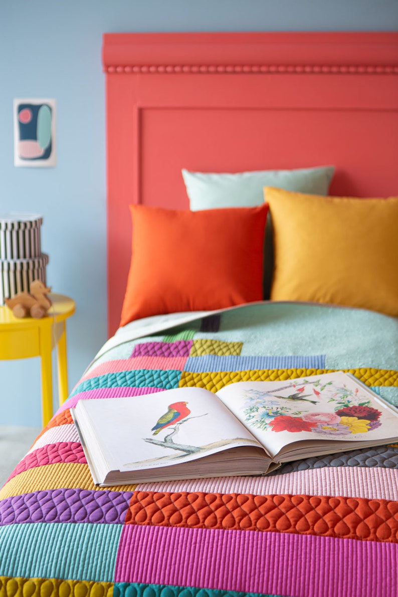 Op de afbeelding: Een kleurrijk patchwork-deken met een open boek erop. Het deken is gemaakt van verschillende gekleurde vierkanten met verschillende texturen. Het boek heeft een afbeelding van een vogel op de omslag.