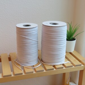 Puede incluir: Dos carretes de cord&oacute;n el&aacute;stico blanco en un estante de madera. Los carretes est&aacute;n envueltos en papel blanco y tienen un cord&oacute;n el&aacute;stico blanco enrollado alrededor de ellos.